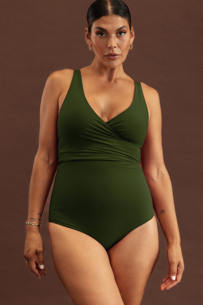 A woman wearing a deep green bikini set, with the high waisted bottoms and bottom seam of the bikini top joining to create the illusion of a one piece swimsuit.