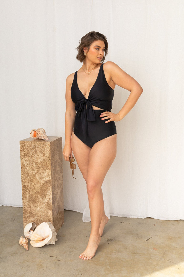 Woman wears an all black, sculpting bikini set standing next to a marbled plinth.
