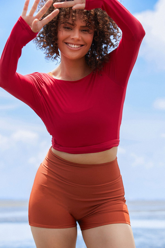 Woman wearing a raspberry coloured long sleeve wrap top with high waisted clay swim shorts.